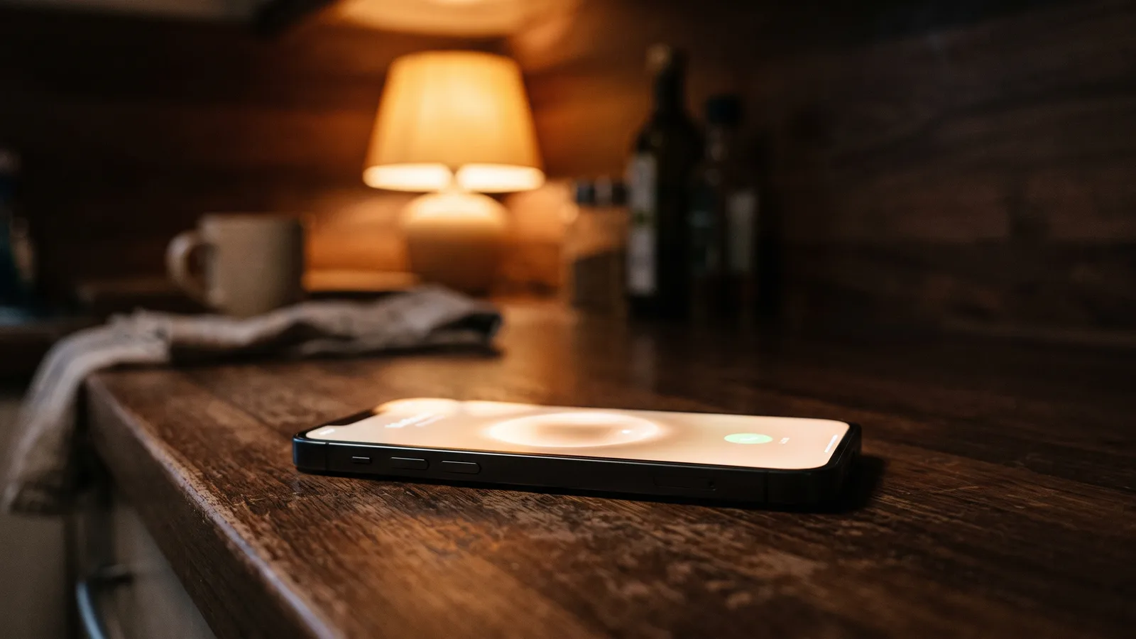 Smartphone lighting up with an incoming call on a kitchen counter at night beside a softly glowing warm reading lamp.